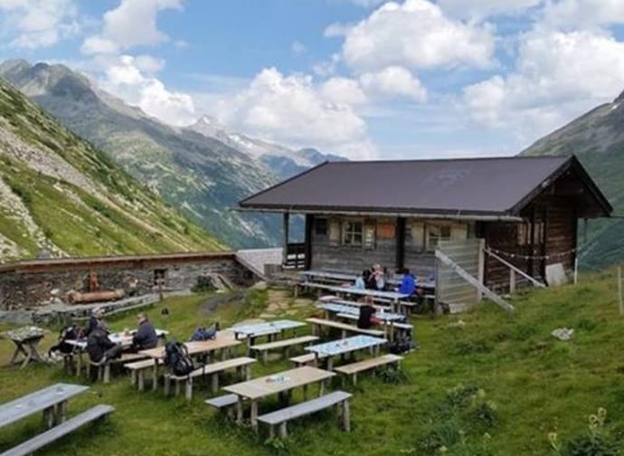 A mountain cabin with outdoor tables, surrounded by green hills and mountains under a blue sky.