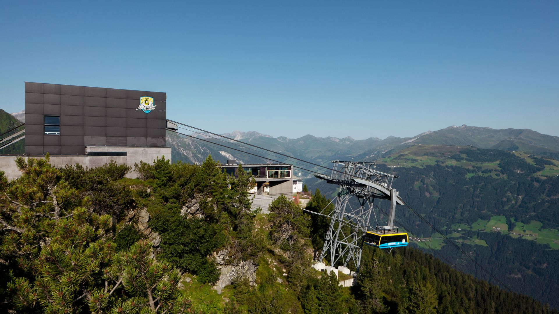 Summertime mountain station of the Ahornbahn cable car by Zillertaler Seilbahnen in Mayrhofen. Modern station with glass façade on a green mountain ridge. Cable car with panoramic view of surrounding Alpine peaks under a clear blue sky.