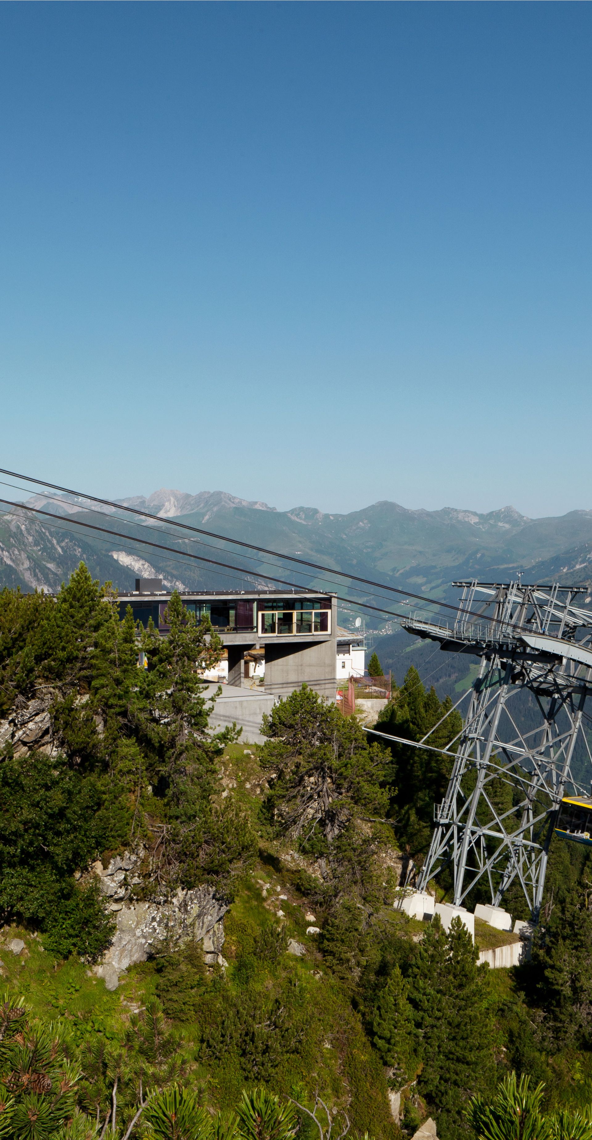 Summertime mountain station of the Ahornbahn cable car by Zillertaler Seilbahnen in Mayrhofen. Modern station with glass façade on a green mountain ridge. Cable car with panoramic view of surrounding Alpine peaks under a clear blue sky.