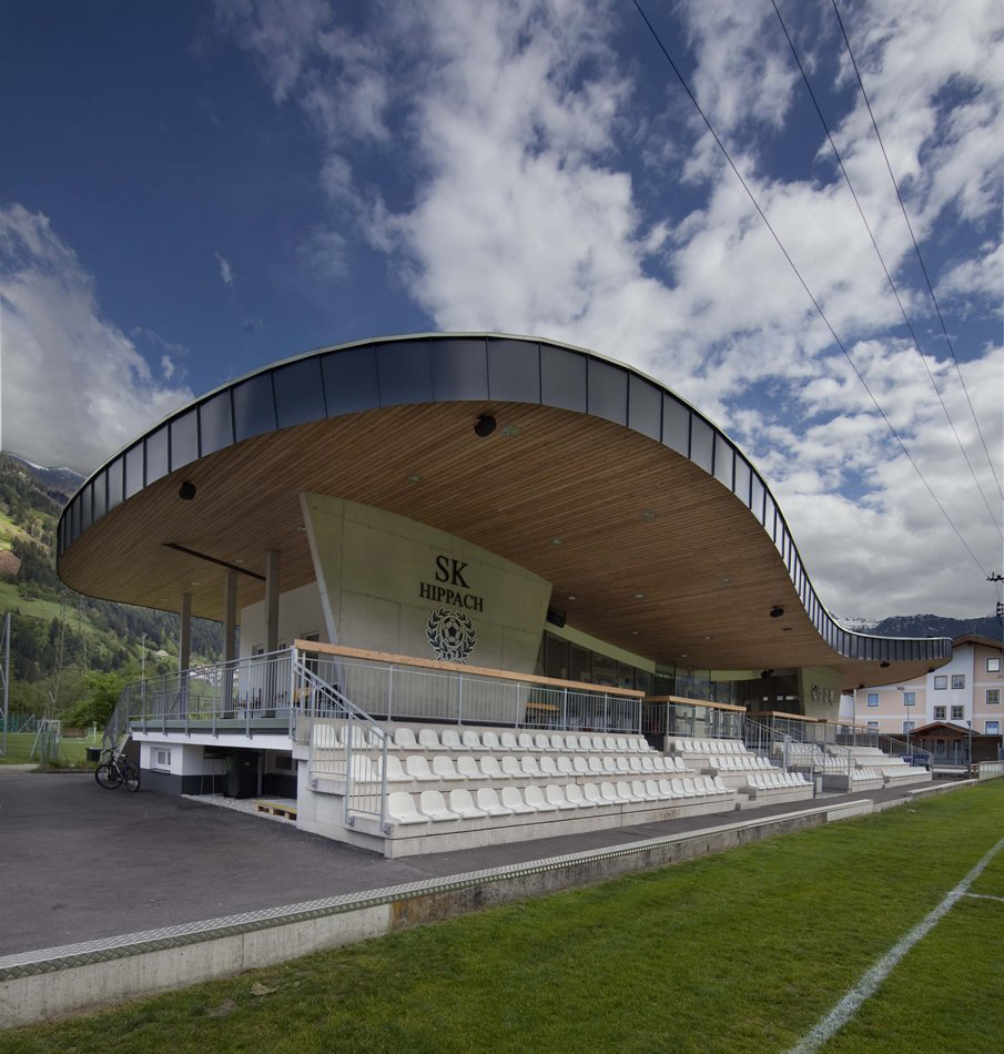 Clubhouse of SK Raika Hippach at Lindenstadion with curved timber roof design, seating area, and mountain backdrop under a blue sky in Tyrol.