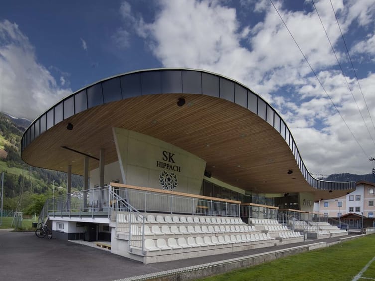 Clubhouse of SK Raika Hippach at Lindenstadion with curved timber roof design, seating area, and mountain backdrop under a blue sky in Tyrol.