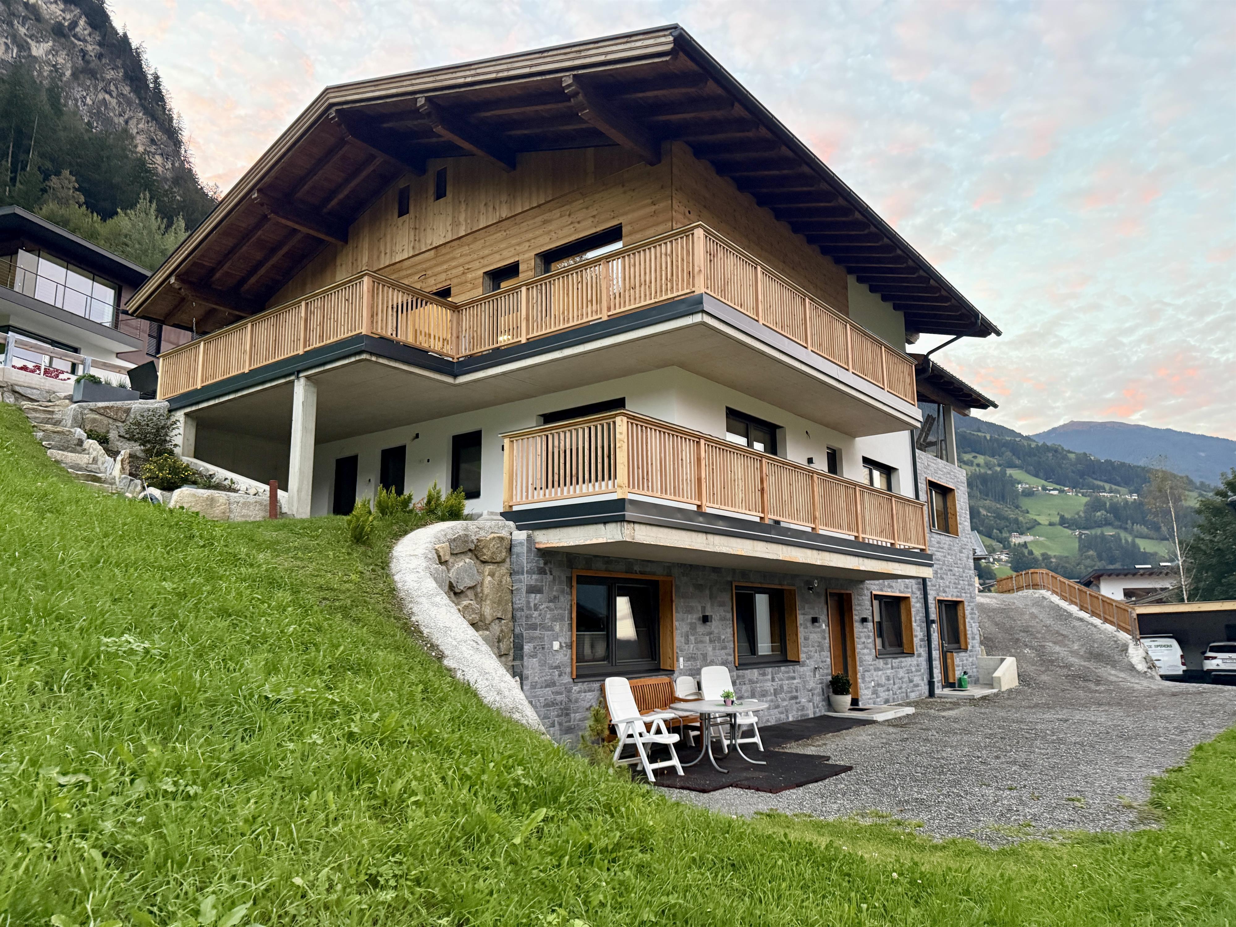Modern house with wood and stone facade, balcony, garden, in mountain surroundings at sunset.