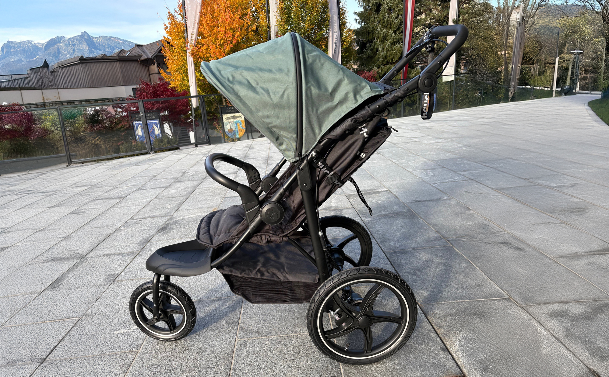 Ein schwarzer Kinderwagen mit grünem Verdeck steht auf einem breiten, glatten Gehweg vor einem modernen Gebäude in Mayrhofen. Der Zugang ist ebenerdig und gut befahrbar.