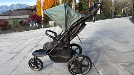 Ein schwarzer Kinderwagen mit grünem Verdeck steht auf einem breiten, glatten Gehweg vor einem modernen Gebäude in Mayrhofen. Der Zugang ist ebenerdig und gut befahrbar.