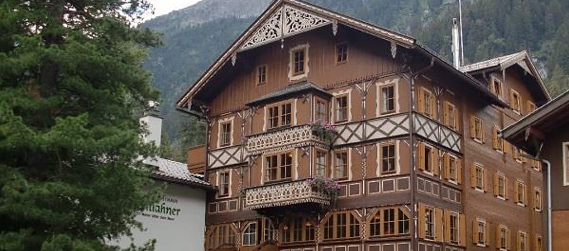 Multi-story wooden house with balcony, stairs, and potted plants in front of mountains.