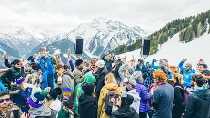 The image shows an outdoor mountain party with people celebrating in the foreground and a DJ in the background.