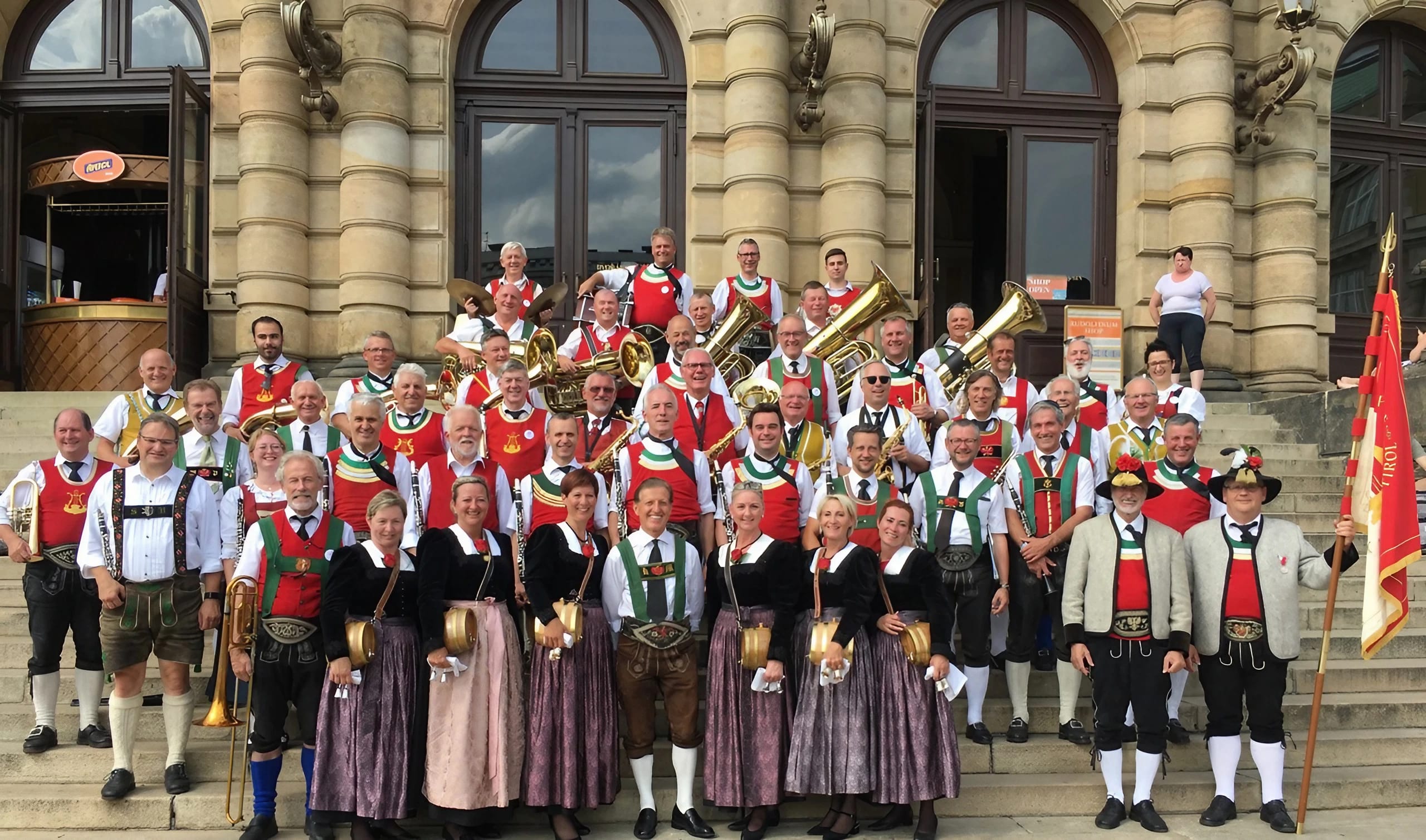 The photo shows a large brass band in traditional costume posing for a group photo in front of a historic building. The musicians are wearing festive hats adorned with feathers, colorful traditional outfits, and are holding various brass instruments.
