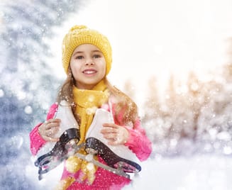 Mädchen mit gelber Mütze hält ein Paar Schlittschuhe vor einer verschneiten Winterlandschaft mit Lichtern. Symbolbild für Eislaufen in Mayrhofen im Zillertal und fröhliche Wintertage mit der Familie.