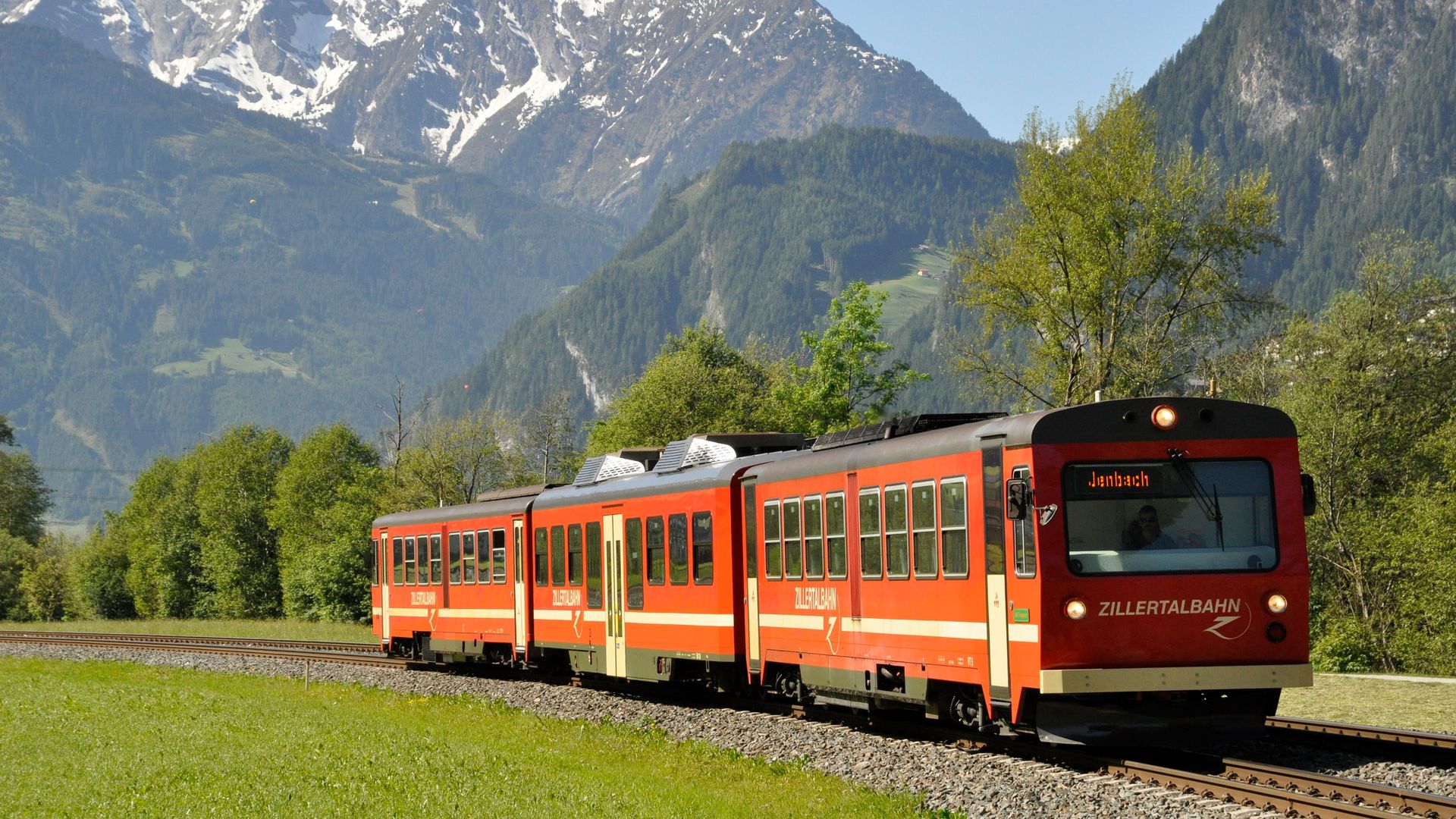 Red and white Zillertalbahn train travels through green alpine meadows with dramatic snow-capped mountain peaks in background. The narrow-gauge railway passes through picturesque spring landscape in the Zillertal valley.