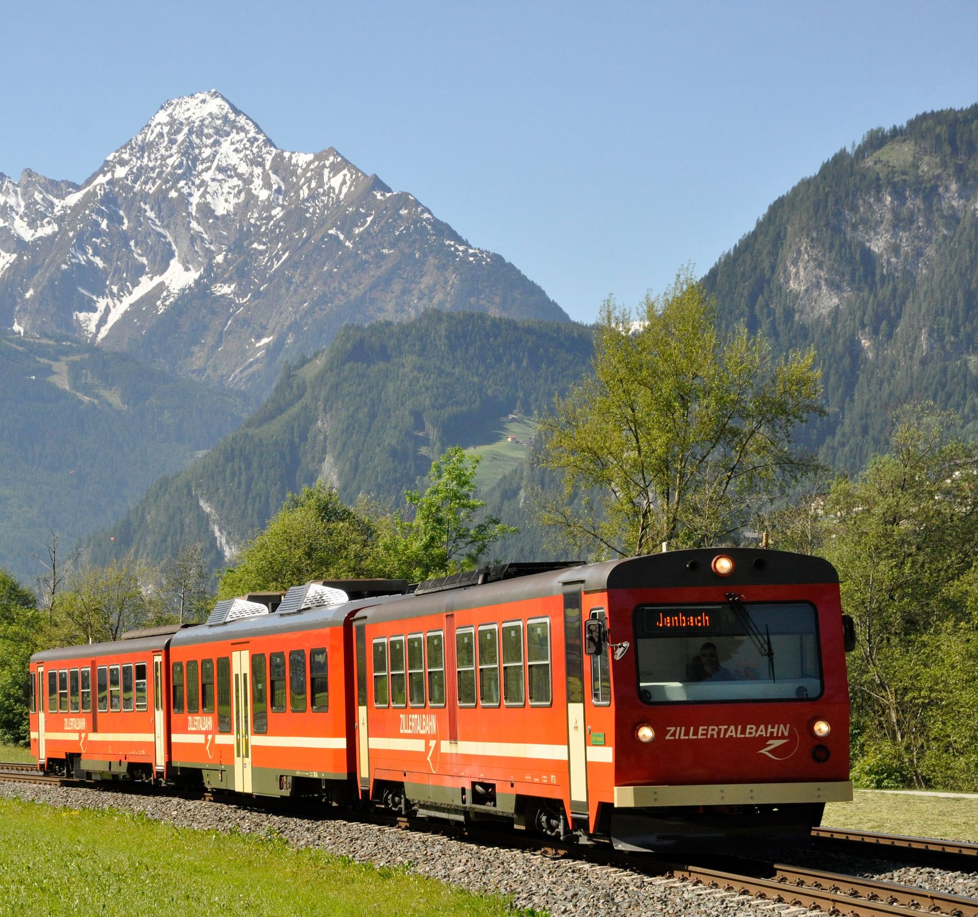 Red and white Zillertalbahn train travels through green alpine meadows with dramatic snow-capped mountain peaks in background. The narrow-gauge railway passes through picturesque spring landscape in the Zillertal valley.