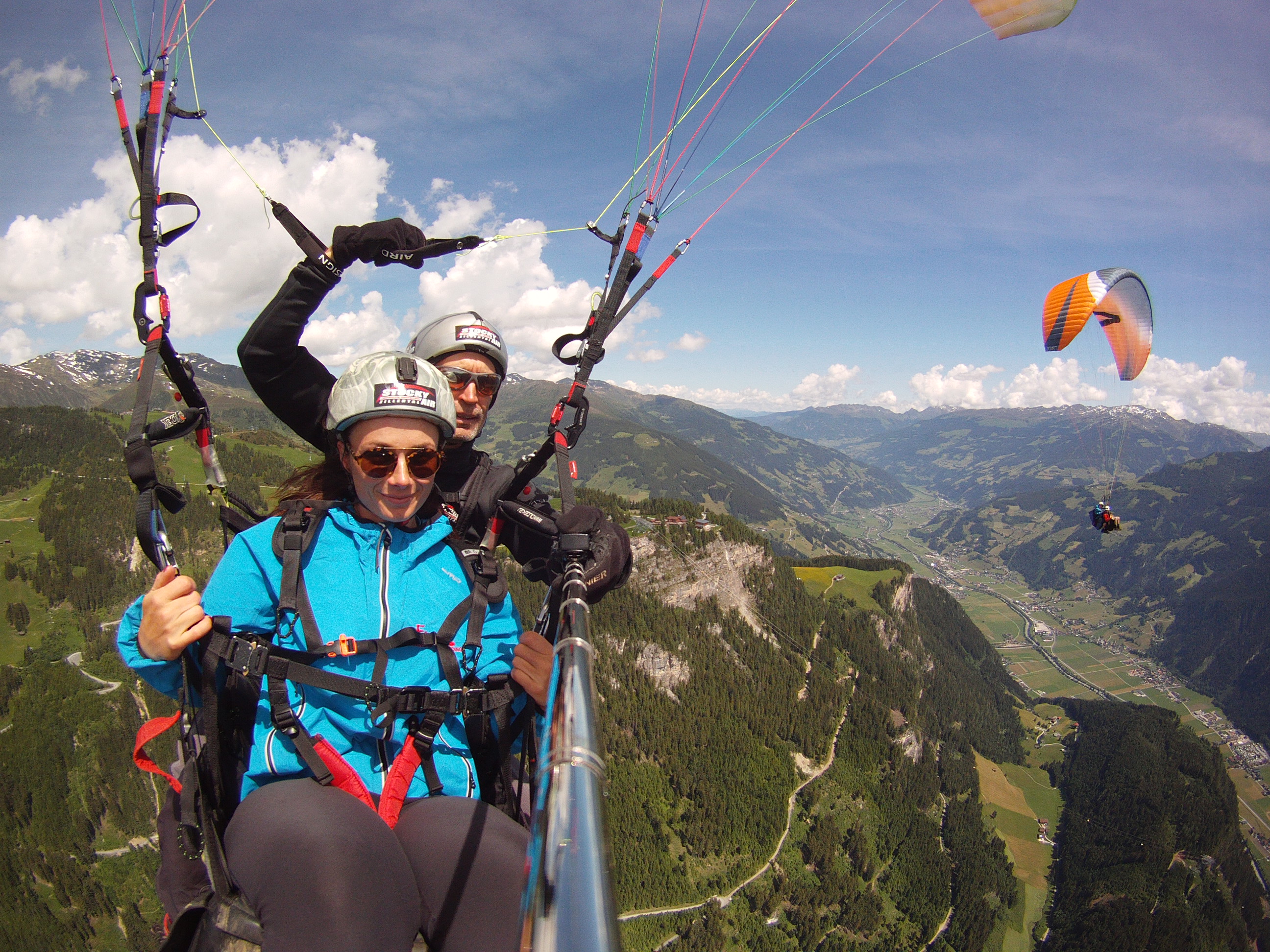 Zwei Personen mit einem Paraglider über einer grünen Landschaft. Im Hintergrund ist ein oranger Paraglider zu sehen.