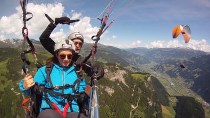 Zwei Personen mit einem Paraglider über einer grünen Landschaft. Im Hintergrund ist ein oranger Paraglider zu sehen.