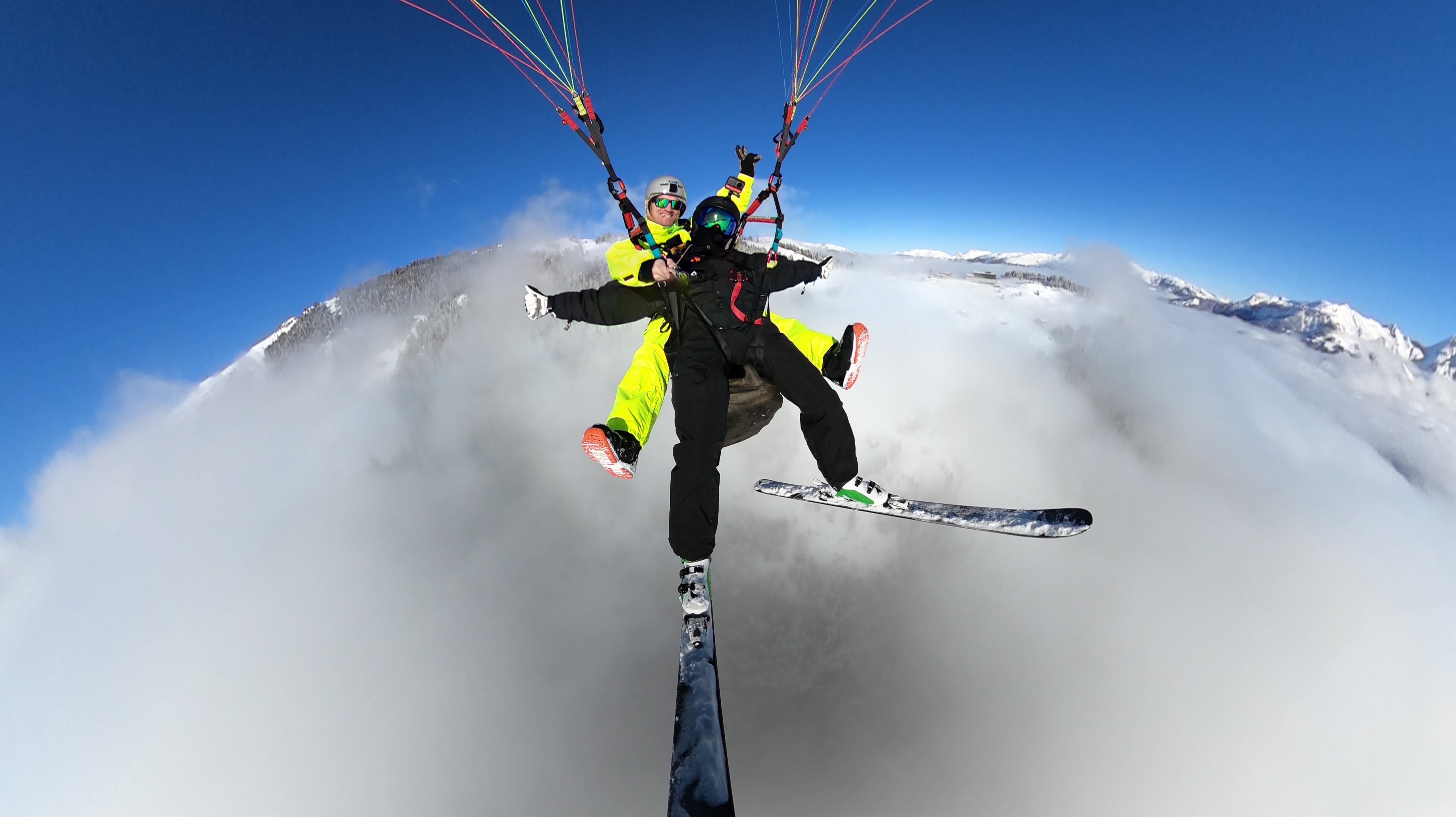The picture shows two people paragliding high above the clouds. Both are wearing weatherproof clothing and helmets, enjoying the breathtaking view together. The sky is bright blue, and white clouds pass beneath them.
