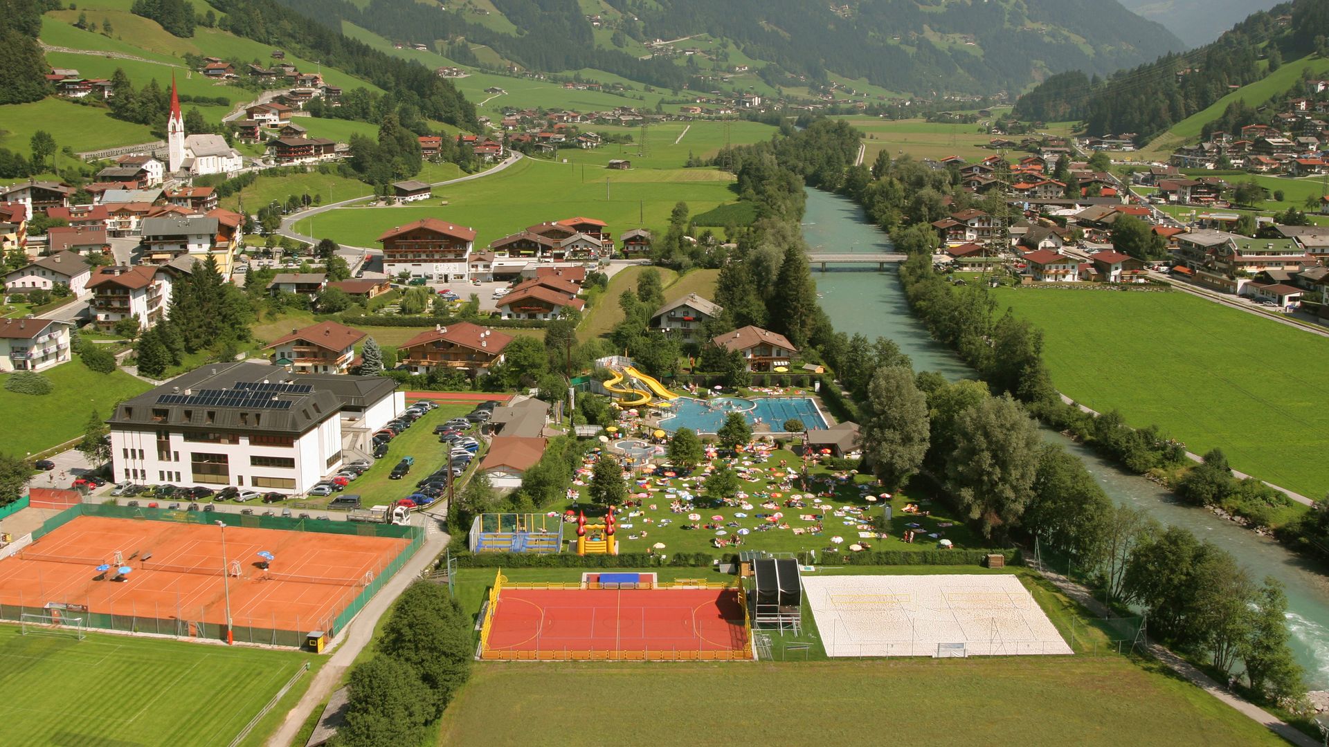 Aerial view of the Summerworld Hippach with outdoor pool, sports courts, and surrounding mountain landscape in Zillertal on a sunny summer day.