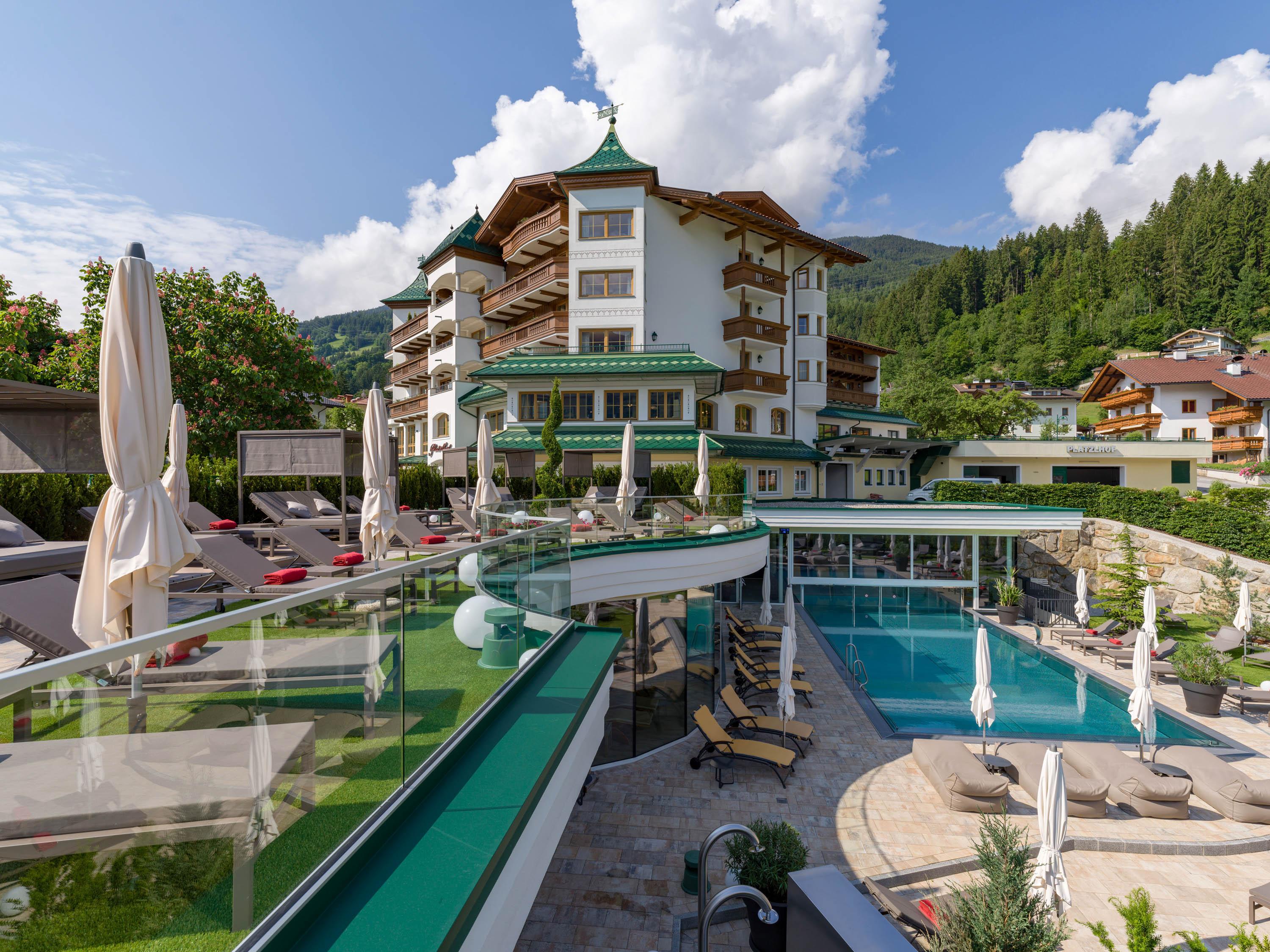 Ein Hotel mit mehreren Balkonen, umgeben von Bergen und Bäumen, mit Pool und Liegestühlen im Vordergrund.