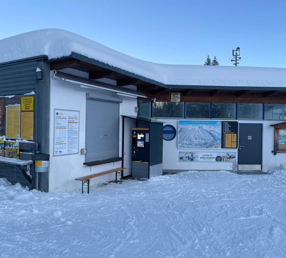 Snow-covered building with info signs, vending machines, and a bench in snow, blue sky in background.