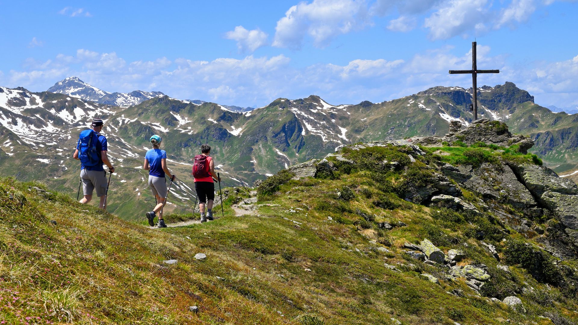Three people are hiking up a green mountainside. They are wearing sportswear and carrying rucksacks. On the right is a large wooden cross on a hill. Rocky mountains with snowfields can be seen in the background under a blue sky with a few clouds.