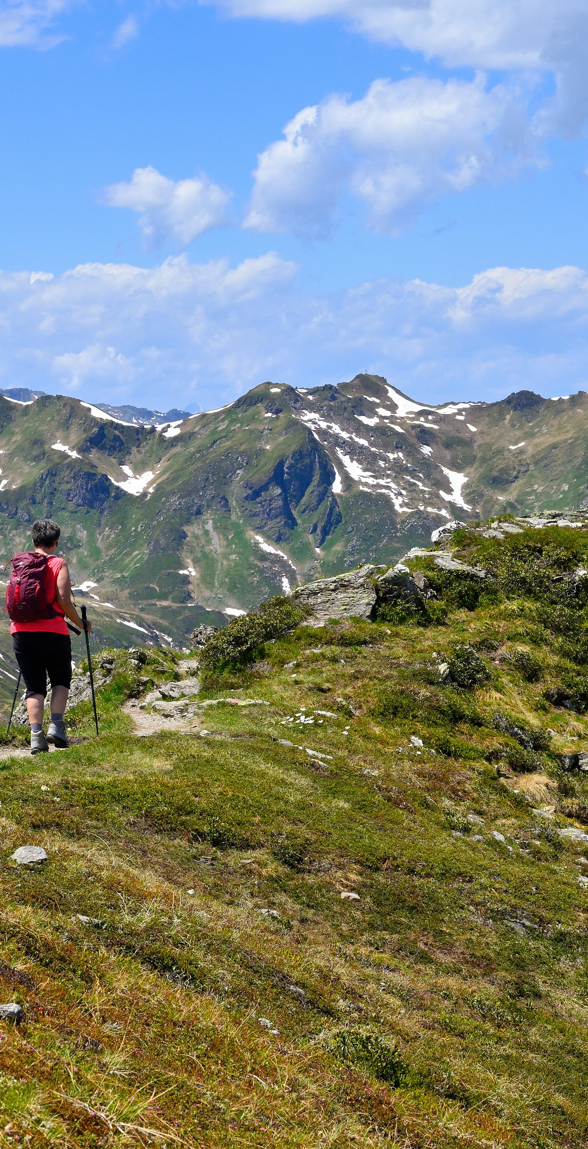 Three people are hiking up a green mountainside. They are wearing sportswear and carrying rucksacks. On the right is a large wooden cross on a hill. Rocky mountains with snowfields can be seen in the background under a blue sky with a few clouds.