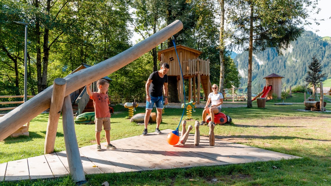 Eine Familie genießt den naturnahen Spielplatz Auenland Sidan in der Ferienregion Mayrhofen-Hippach – mit Holzspielgeräten, Baumhäusern und viel Platz zum Spielen und Entspannen im Grünen, umgeben von Bäumen und Bergen.