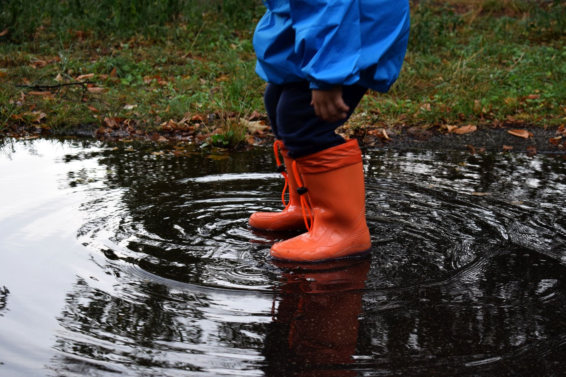 A child wearing a blue rain jacket and orange rubber boots stands in a puddle. The water ripples around their feet. The ground is wet, covered with grass and fallen leaves. The scene feels autumnal and rainy.