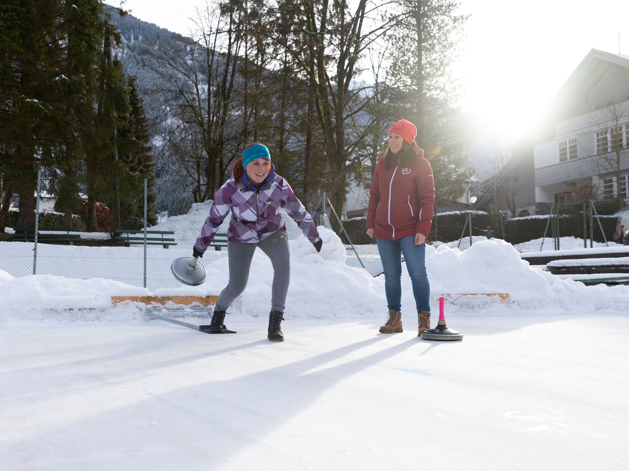 Zwei Personen beim Eisstockschießen auf dem Eislaufplatz Mayrhofen im Zillertal. Auf der sonnigen Eisfläche vor verschneiten Bergen genießen sie gemeinsam den winterlichen Freizeitspaß.