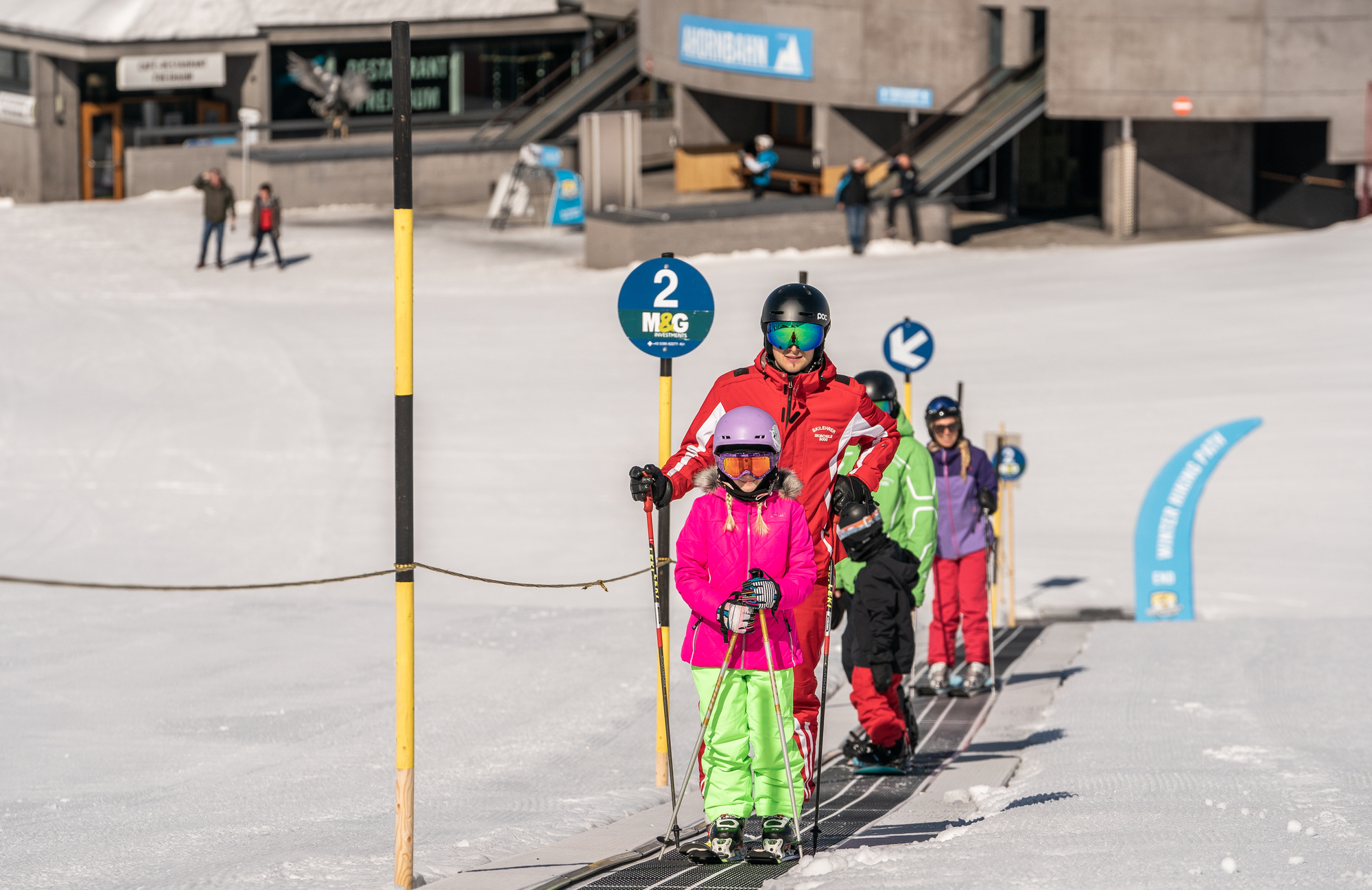 Children taking skiing lessons in Mayrhofen, instructed by ski instructors on a conveyor belt lift, all wearing colorful ski clothing and helmets.