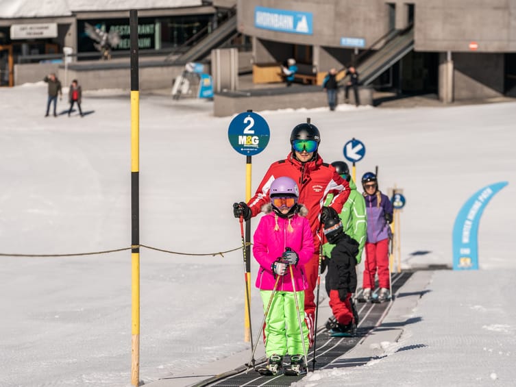 Children taking skiing lessons in Mayrhofen, instructed by ski instructors on a conveyor belt lift, all wearing colorful ski clothing and helmets.