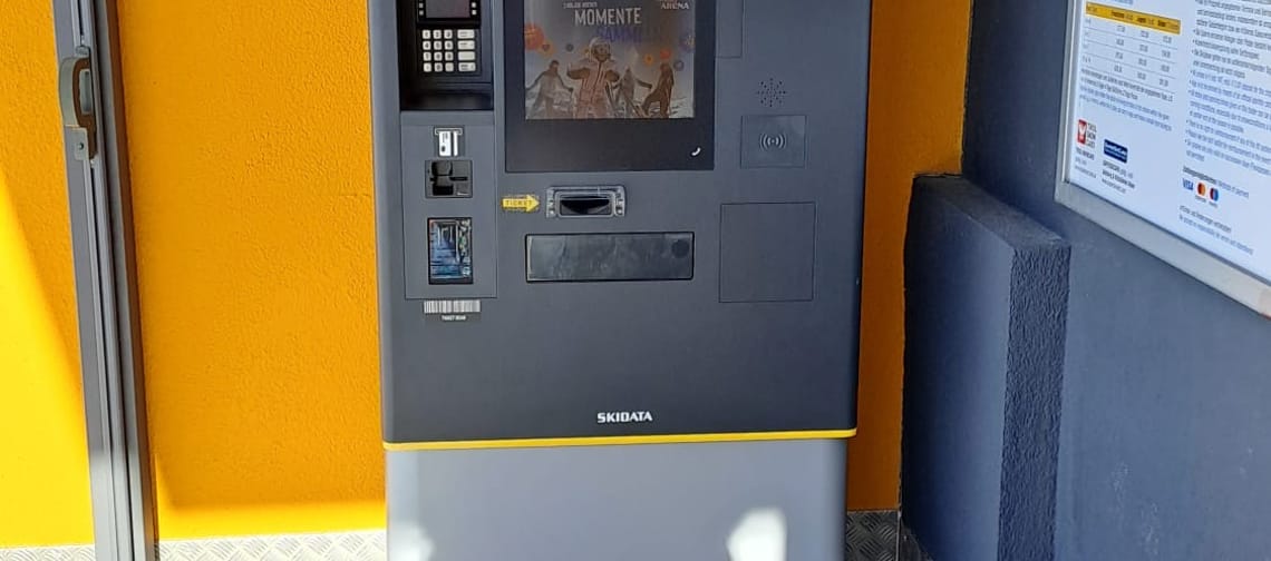 Ticket kiosk against an orange wall on wooden planks with snow patches in front.
