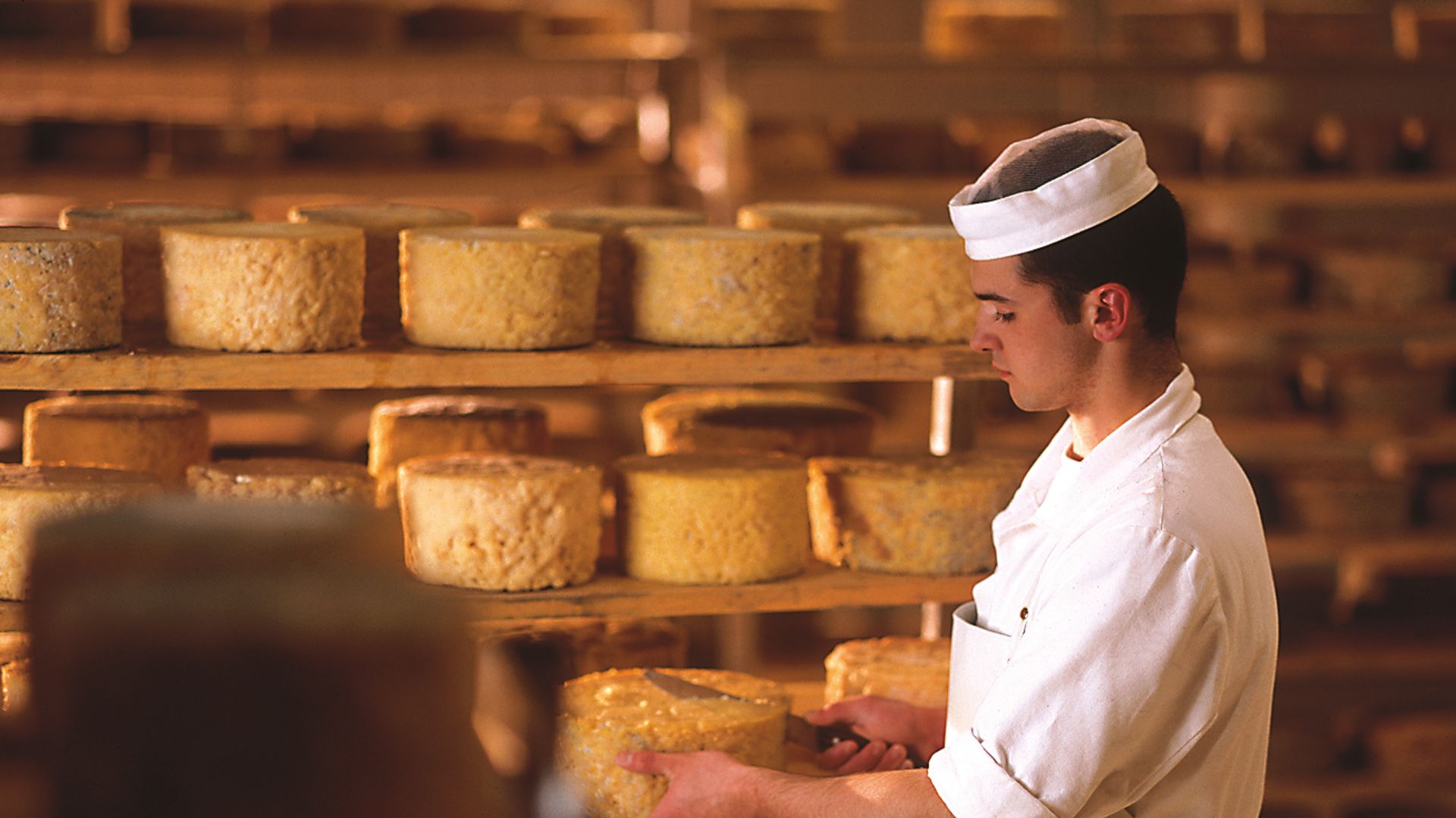 Mann in weißer Arbeitskleidung prüft runde Käselaibe in einem Reiferaum mit Holzregalen in der Erlebnissennerei Zillertal in Mayrhofen. Viele gelbe Käselaibe liegen gestapelt, warme Atmosphäre.