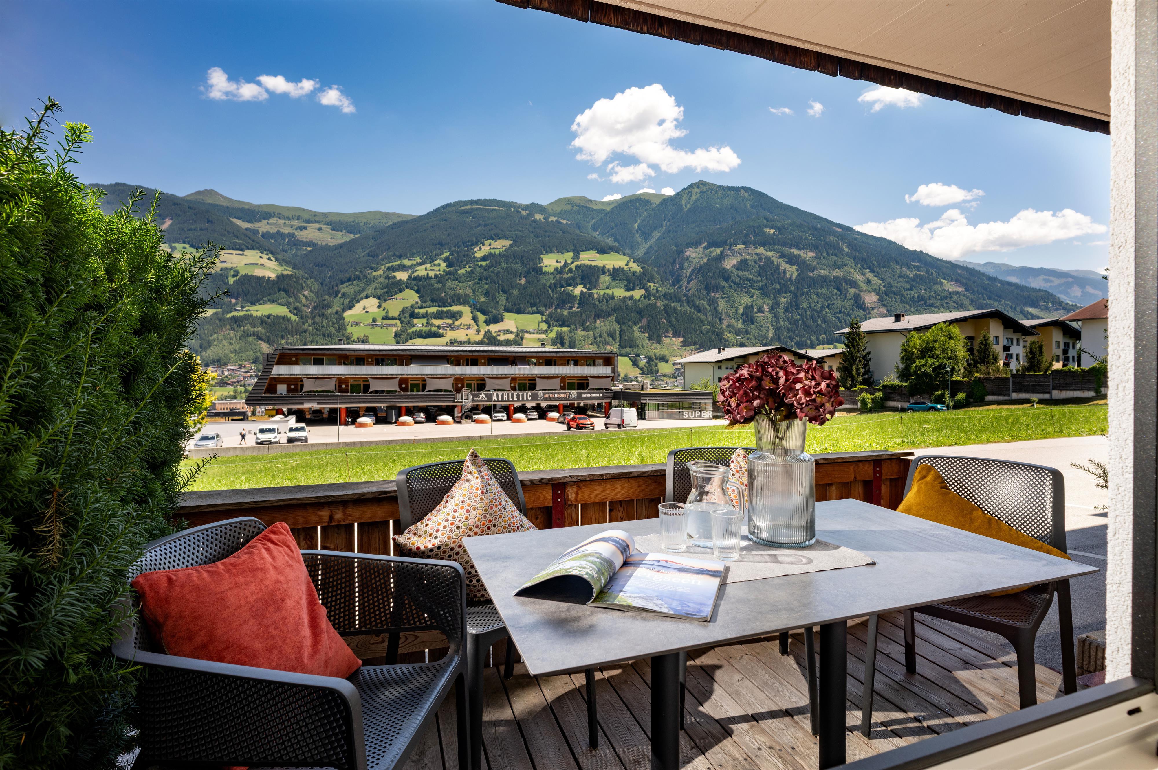 Blick auf eine Terrasse mit Tisch, Stühlen, Kissen und Blick auf Berge und Häuser im Hintergrund bei schönem Wetter.