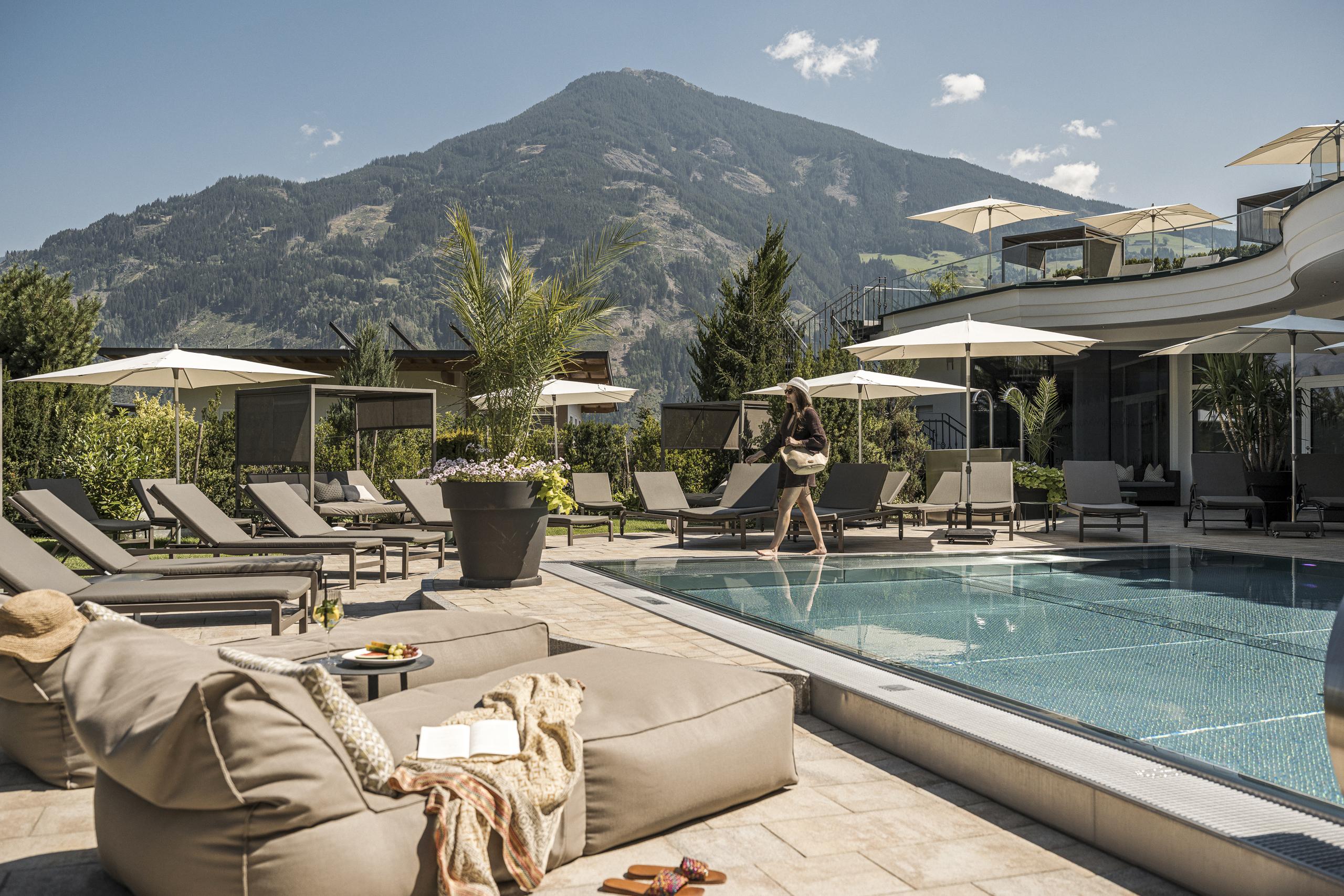 Poolbereich mit Liegen, Sonnenschirmen und einer Frau, die am Poolrand steht, vor Bergen und modernem Gebäude.