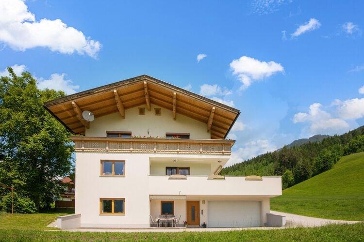 Modernes Haus mit weißer Fassade, Holzdetails und Balkon, vor grüner Wiese und blauer Himmel.