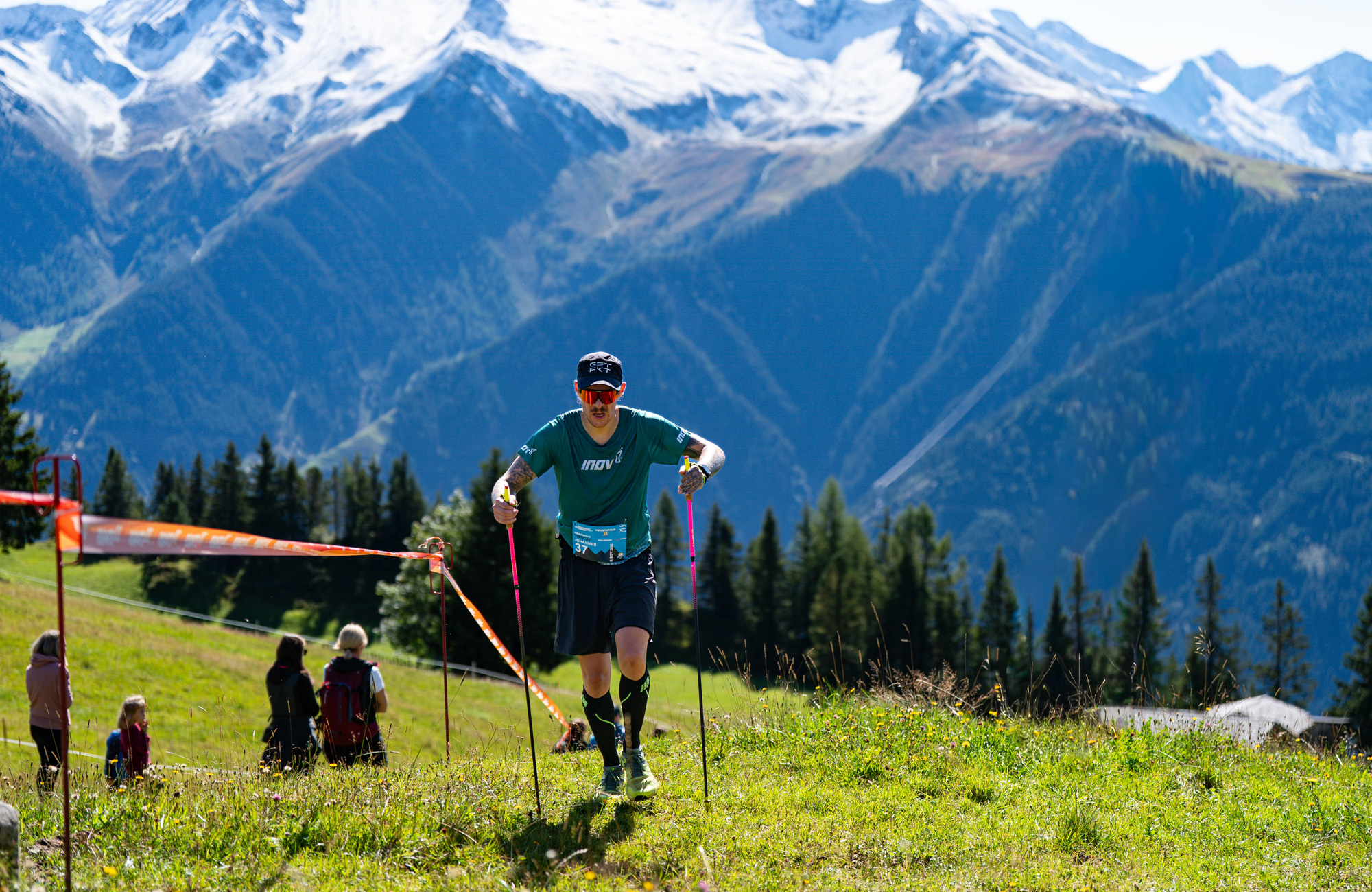 Trail runner competing in the Mayrhofen Ultraks Zillertal on a mountain trail above Mayrhofen, surrounded by alpine meadows, forested slopes and the peaks of the Zillertal Alps.