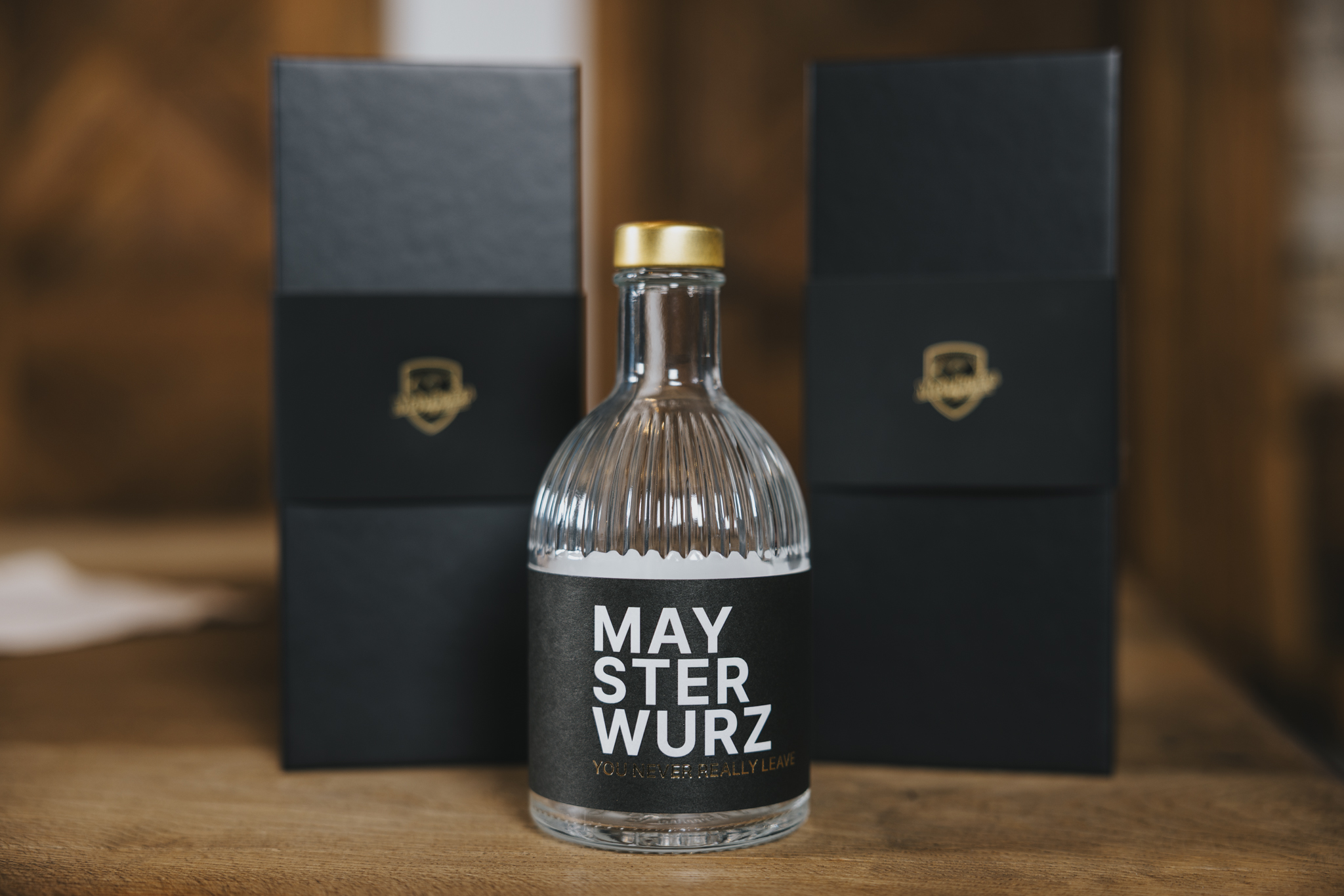 Edelbrand „Maysterwurz“ in einer klaren Glasflasche mit goldenem Verschluss, davor zwei elegante schwarze Verpackungen mit goldfarbenem Logo, auf einem Holztisch platziert.