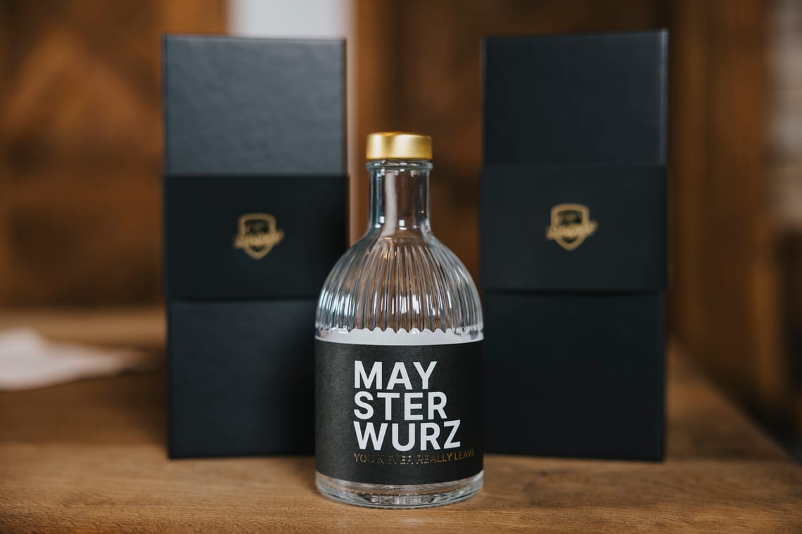 Edelbrand Maysterwurz Edelbrand „Maysterwurz“ in einer klaren Glasflasche mit goldenem Verschluss, davor zwei elegante schwarze Verpackungen mit goldfarbenem Logo, auf einem Holztisch platziert.