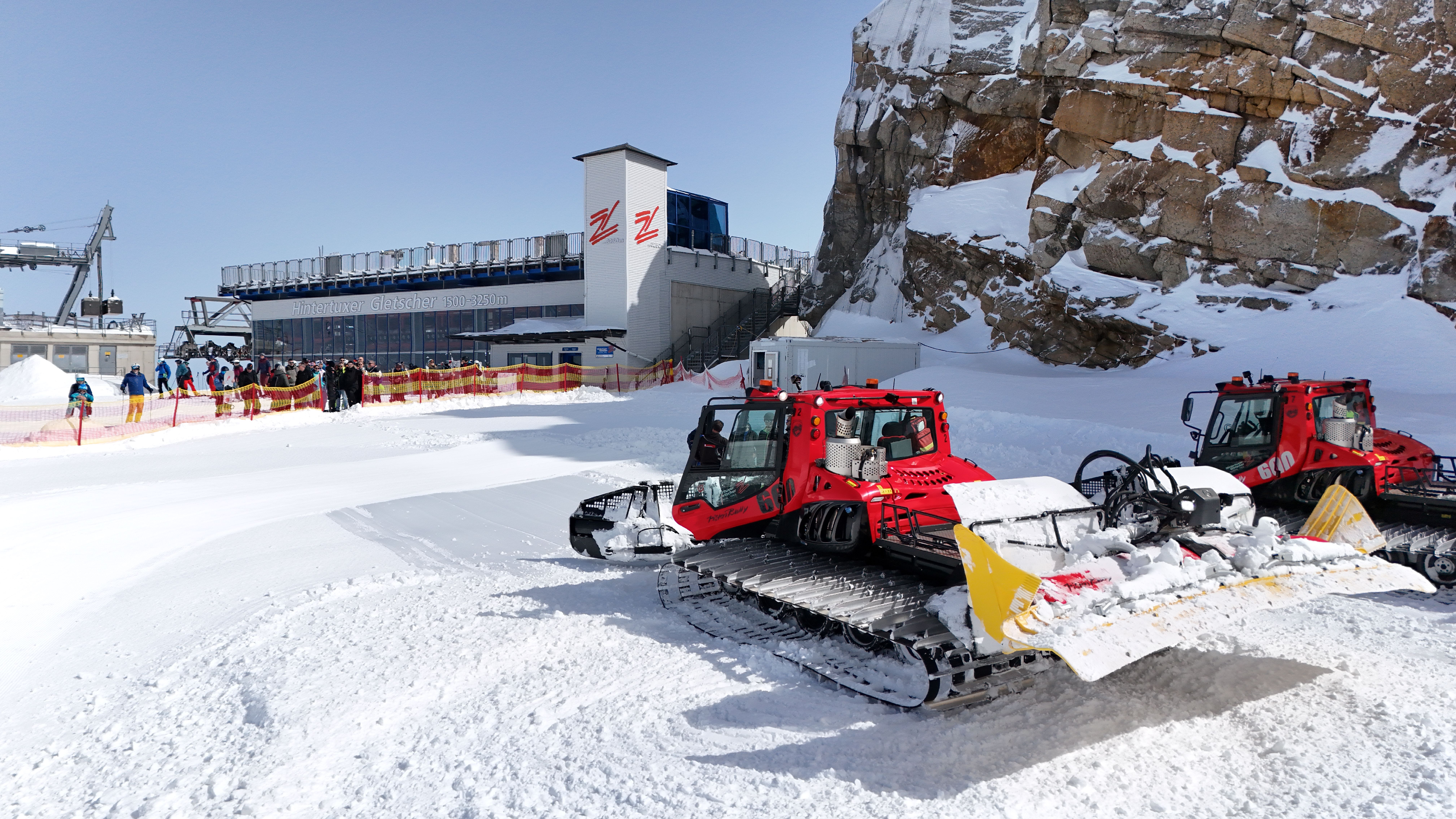 Skiliftstation auf verschneitem Berg mit Schneepflügen und Warteschlange, umgeben von Felsen und Schnee.
