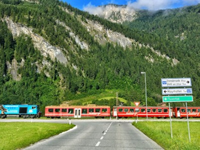 The red Zillertalbahn railway crosses the road at a level crossing in an alpine landscape. Green wooded mountain slopes and rock faces frame the valley. Traffic signs and barriers regulate the intersection between train and car traffic.