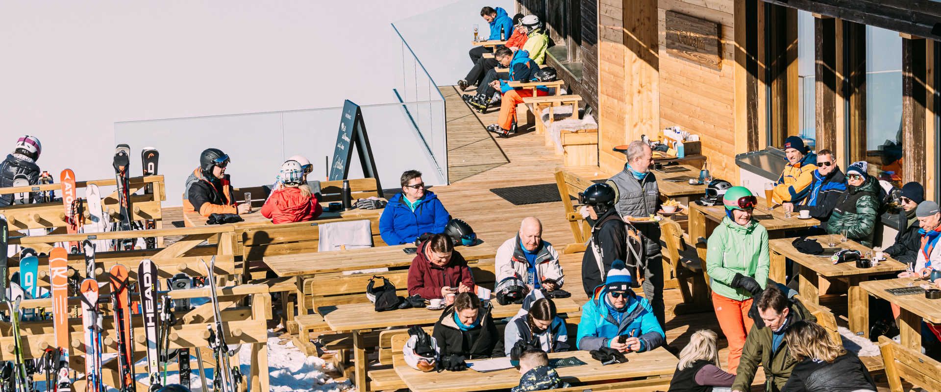 Sonnenterrasse der Goldalm am Sportberg Goldeck mit Gästen, Skiausrüstung und Panoramablick in die winterliche Berglandschaft.