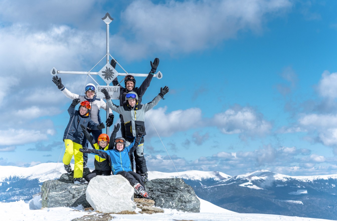 Winter sports enthusiasts at the summit cross of the Klippitztörl in Carinthia, with snow-capped mountains and a sweeping Alpine panorama.