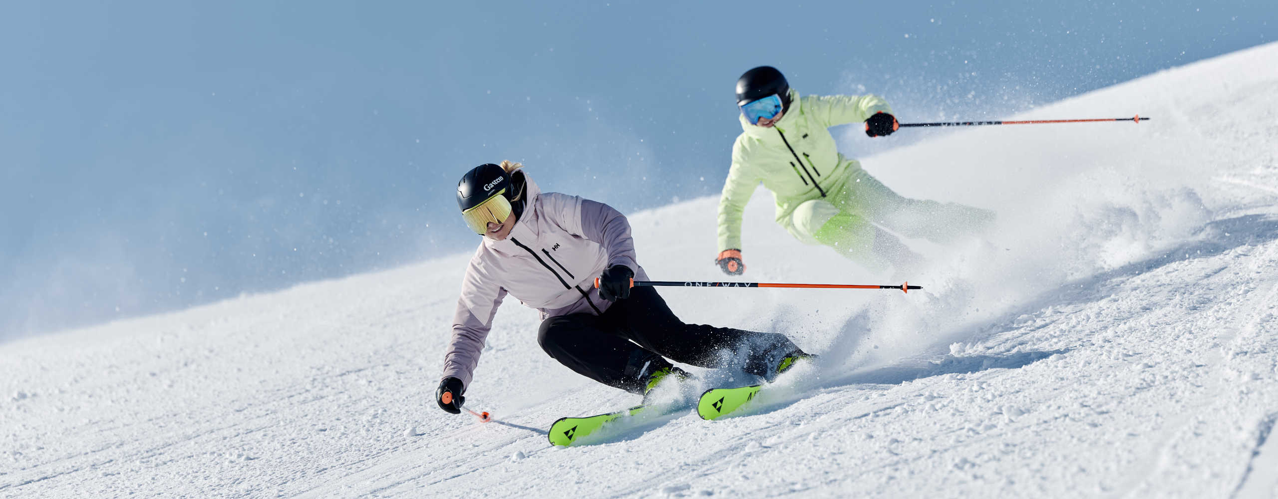 Two skiers carve turns down the freshly groomed slopes of Skigastein under a bright blue sky.