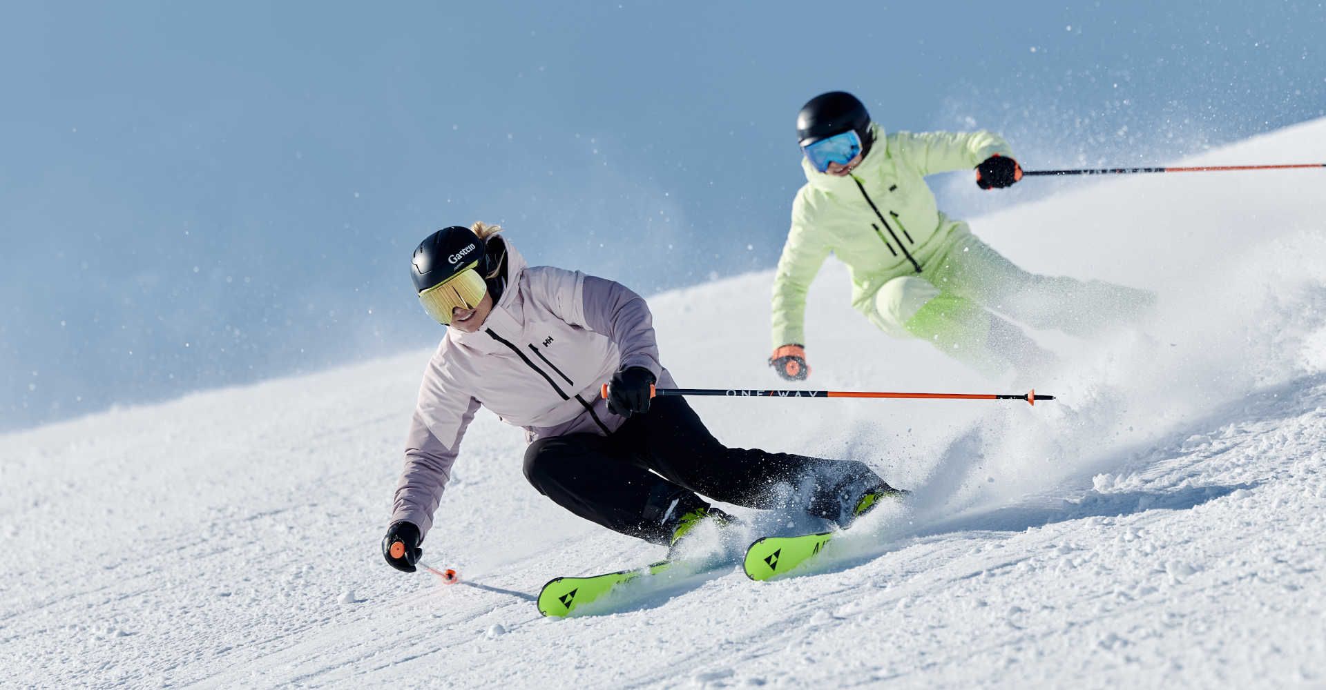 Two skiers carve turns down the freshly groomed slopes of Skigastein under a bright blue sky.