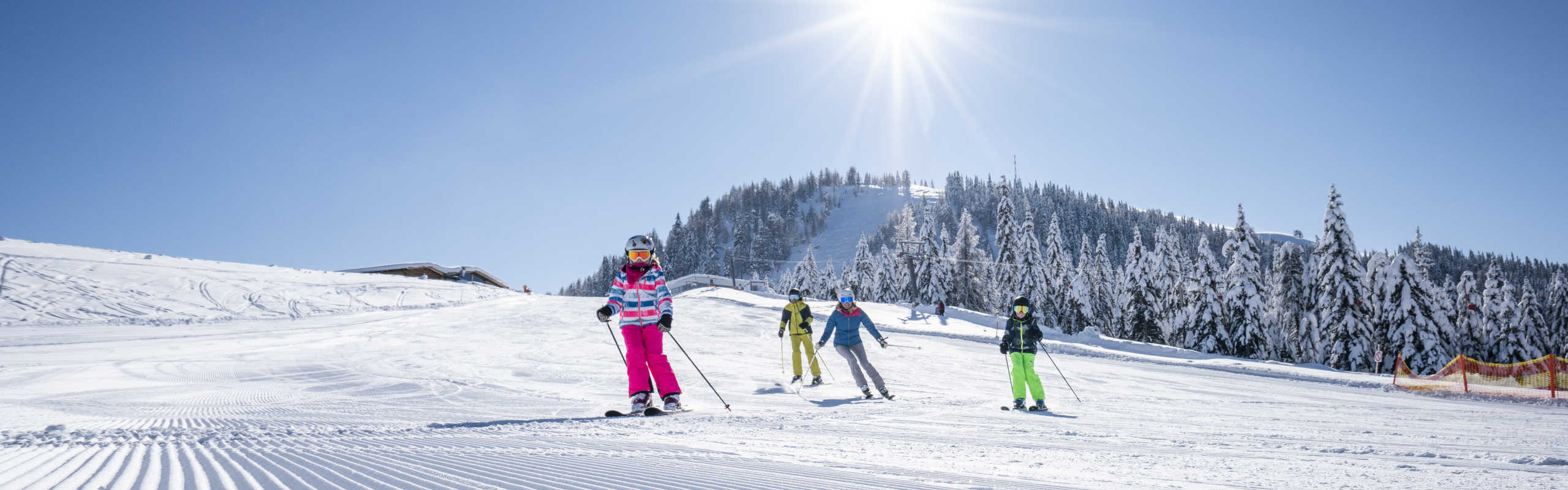 Kinder beim Skifahren am Sportberg Goldeck auf einer sonnigen, verschneiten Piste mit Winterwald im Hintergrund.