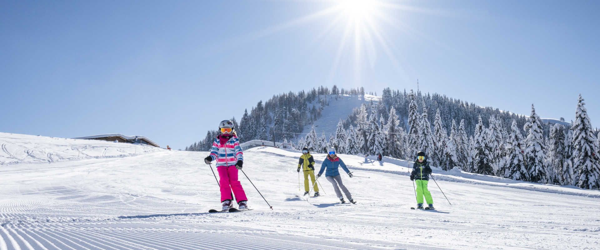 Kinder beim Skifahren am Sportberg Goldeck auf einer sonnigen, verschneiten Piste mit Winterwald im Hintergrund.