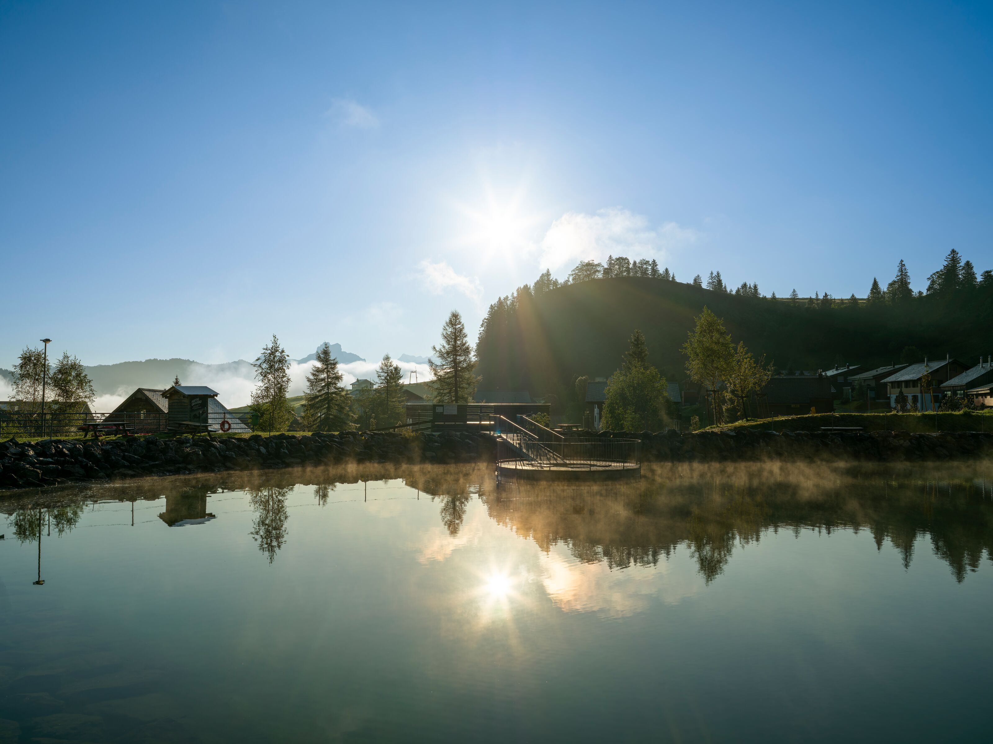 Mountain lake in morning light reflecting the sun, surrounded by houses and alpine landscape.