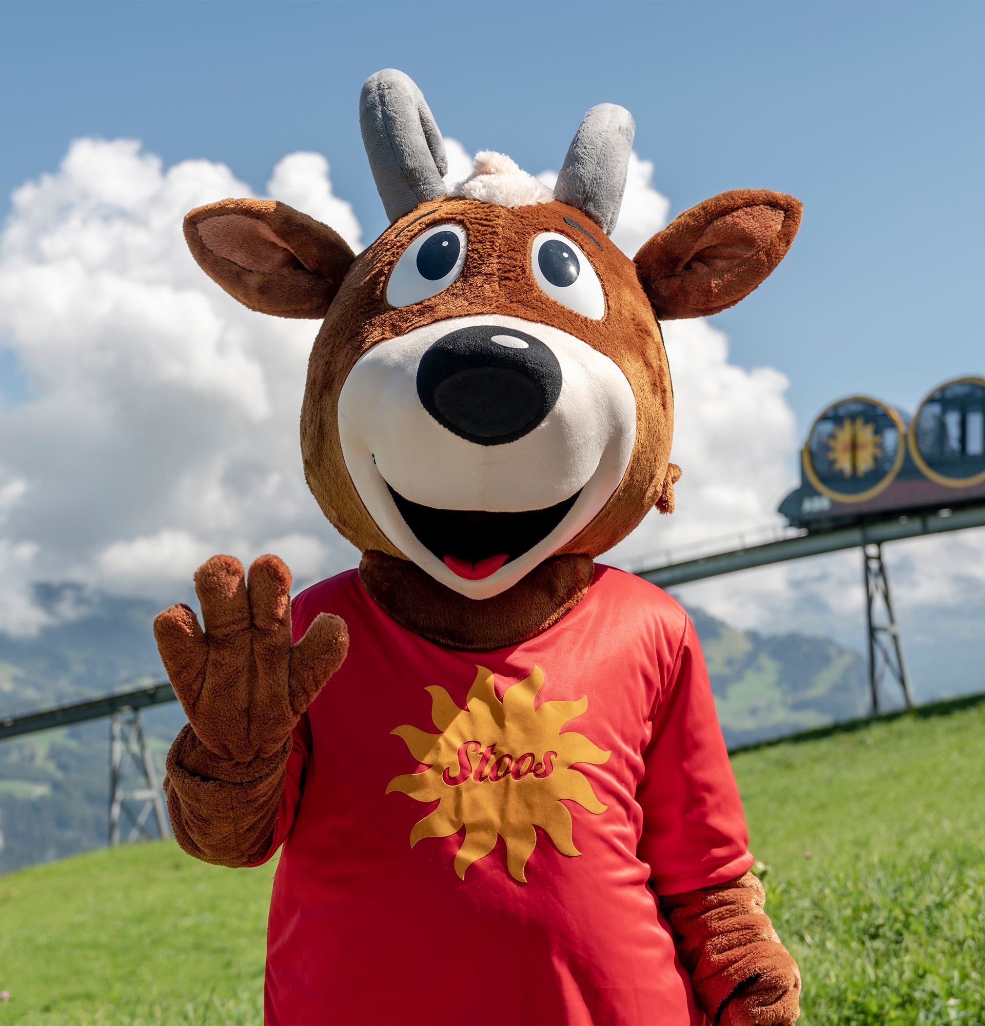 Maskottchen steht auf einer Bergwiese und winkt, im Hintergrund die Stoosbahn und alpine Landschaft.