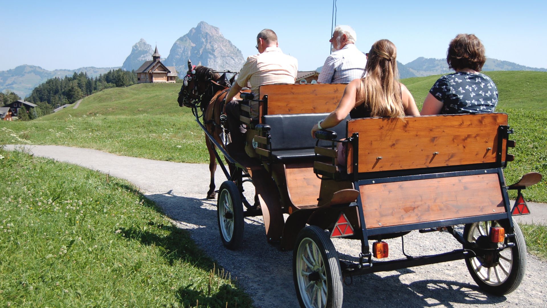 Group riding in a horse-drawn carriage along a mountain path through an alpine landscape.