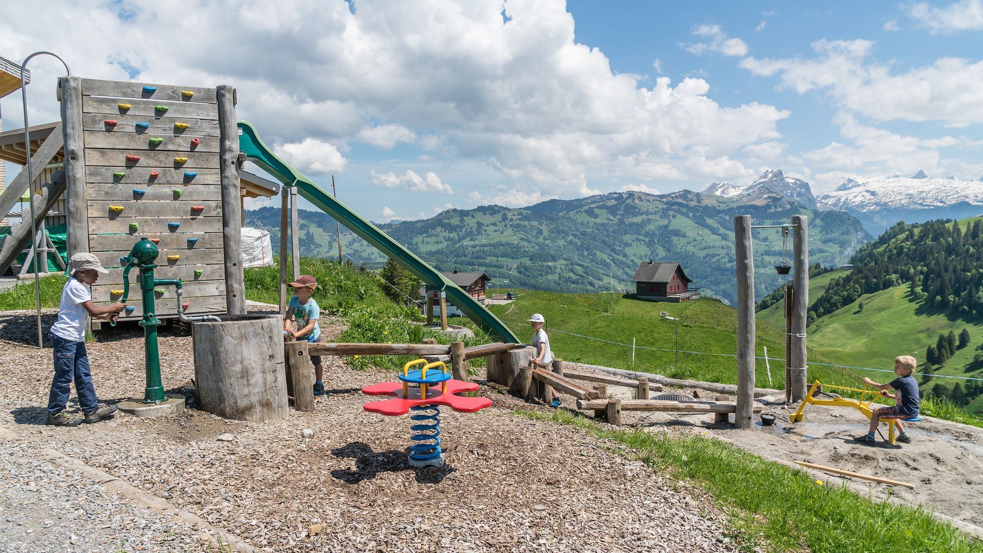 Kinder spielen auf einem Spielplatz mit Kletterwand und Seilbahn in alpiner Umgebung.