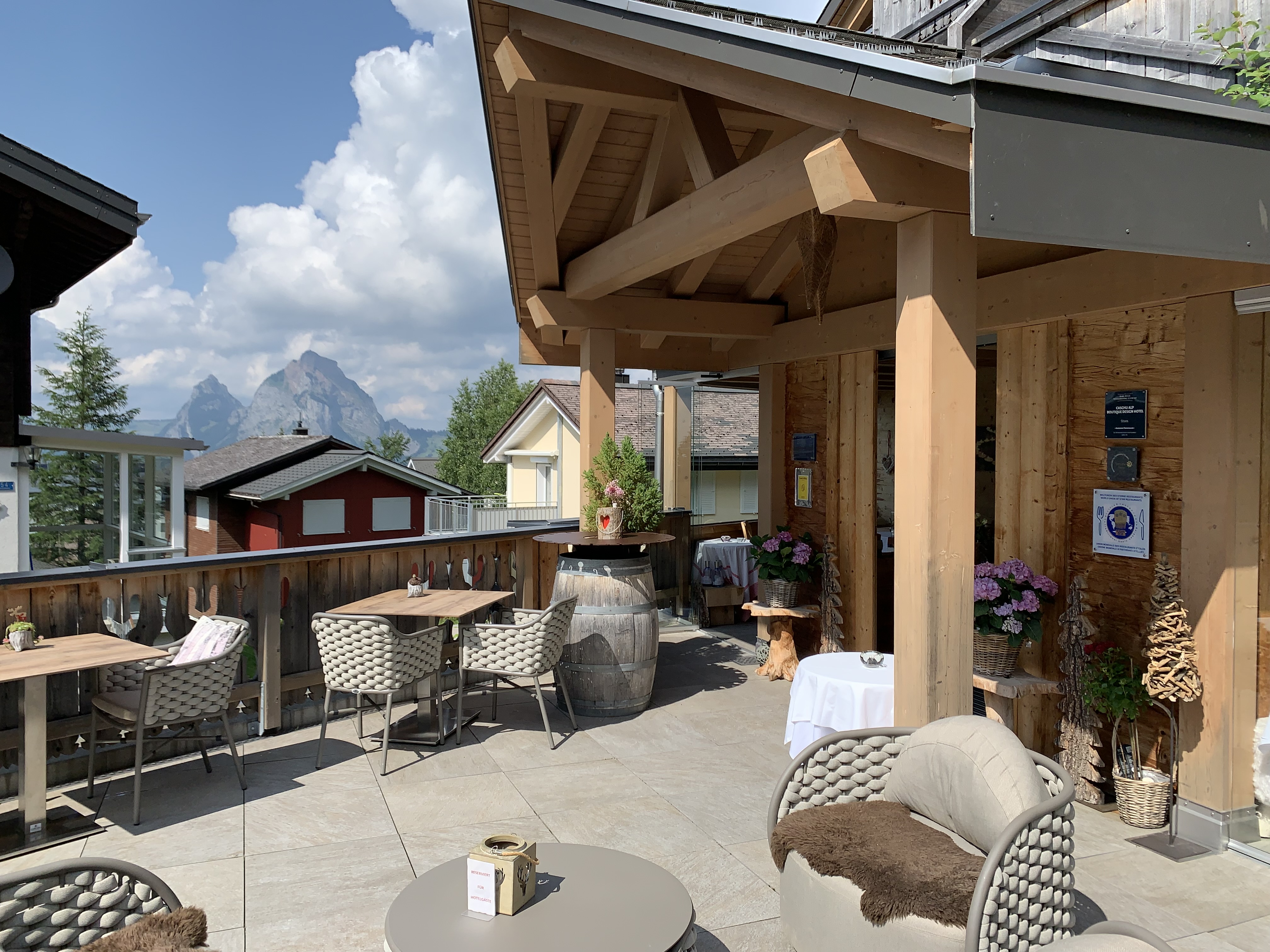 Restaurant terrace with seating and views of buildings and alpine surroundings.