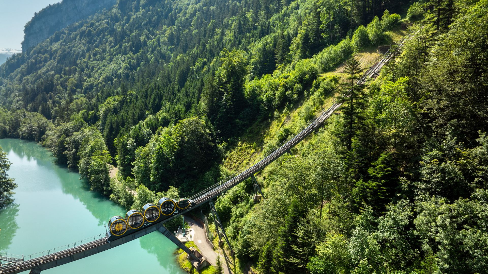 Standseilbahn Schwyz–Stoos auf einer steilen Strecke über einer Brücke entlang eines Sees, umgeben von dichter Bergwaldlandschaft.
