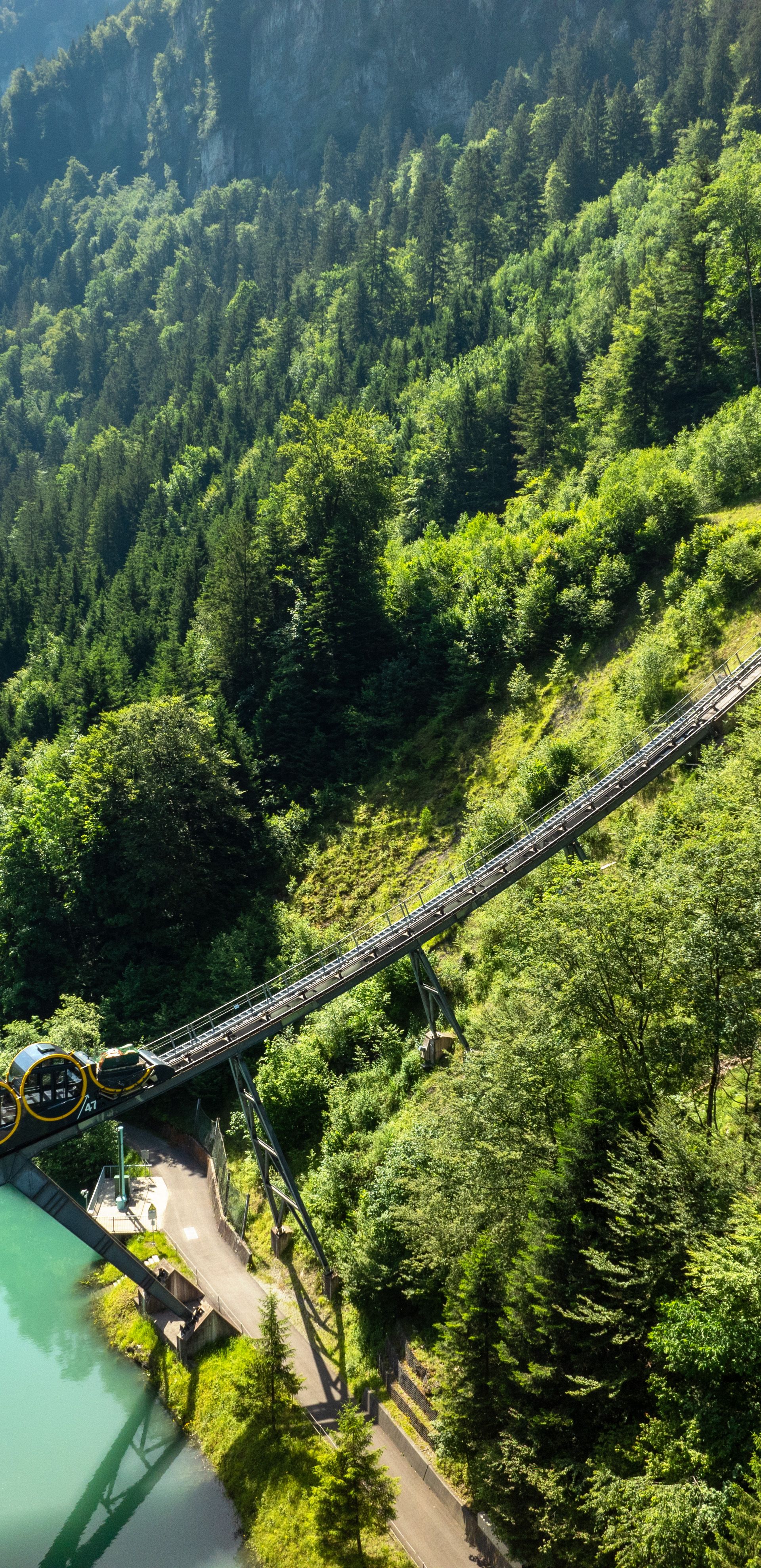 Standseilbahn Schwyz–Stoos auf einer steilen Strecke über einer Brücke entlang eines Sees, umgeben von dichter Bergwaldlandschaft.